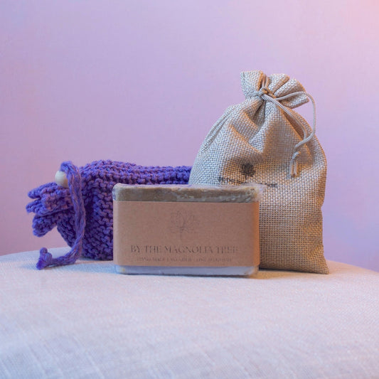A bar of lavender soap sits on a light fabric surface, styled with a small beige drawstring pouch of lavender and a purple knitted wash mitt in a soft pink-lit setting.