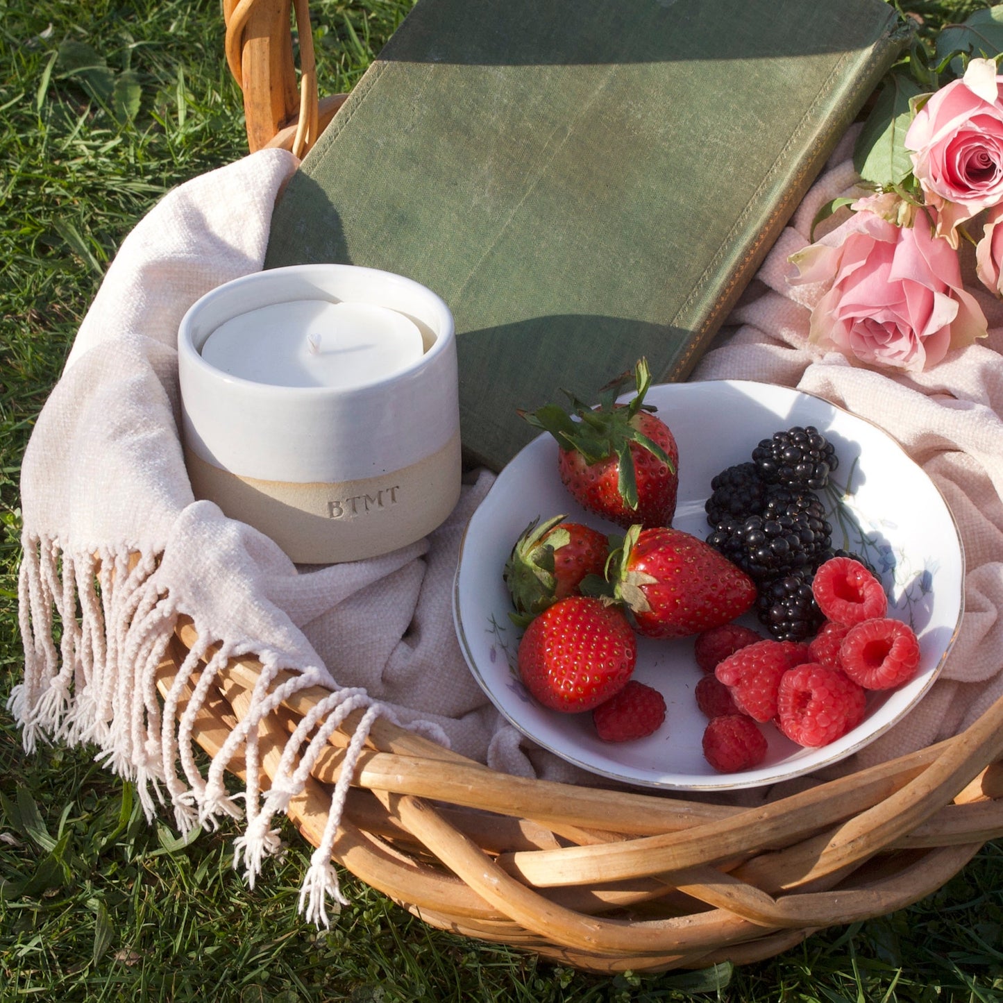 Picnic basket with a bowl of berries, a book, and a white container on a grassy surface.