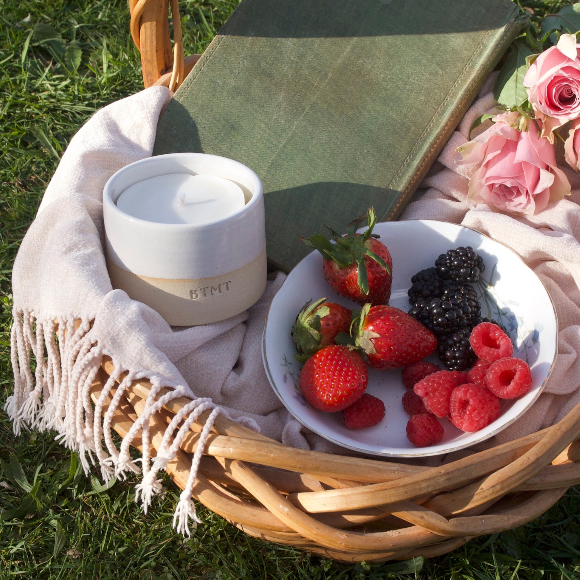 Picnic basket with a bowl of berries, a book, and a white container on a grassy surface.