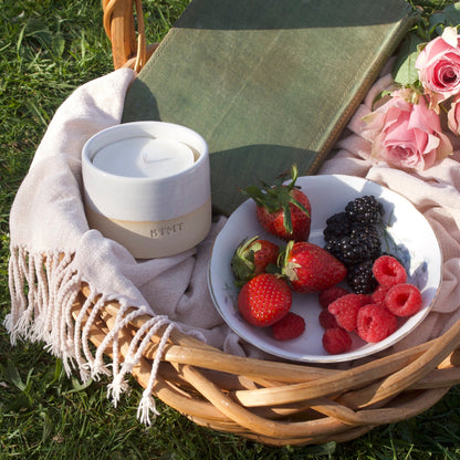 Picnic basket with a bowl of berries, a book, and a white container on a grassy surface.